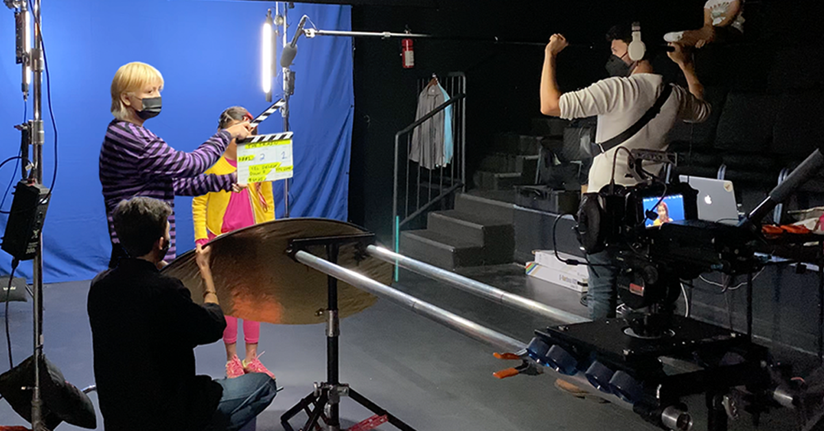Crew members are ready to begin filming a scene. One member holds a reflector, one the camera and another the sound equipment. A young actress stands in front a blue screen, while a student holds the slate in front of her.