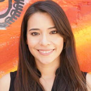 Headshot of a female with long medium brown hair in a black shirt. Behind her is a background painted with warm tones in a gradient pattern.