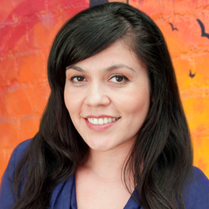 Headshot of a female with dark brown hair and a blue shirt. Behind her is a background painted with warm tones in a gradient pattern.