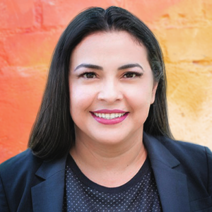 Headshot of a female with long black hair in a black blazer and polka dotted shirt. Behind her is a background painted with warm tones in a gradient pattern.