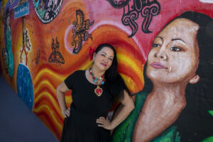 Josefina Lopez posing in front of a colorful mural that includes a painted version of herself.