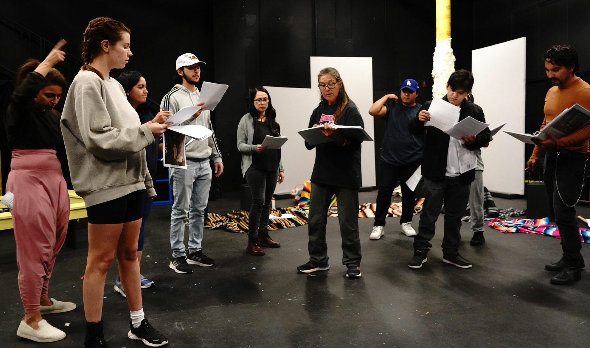A group of actors standing around in a semi circle, rehearse while holding scripts.