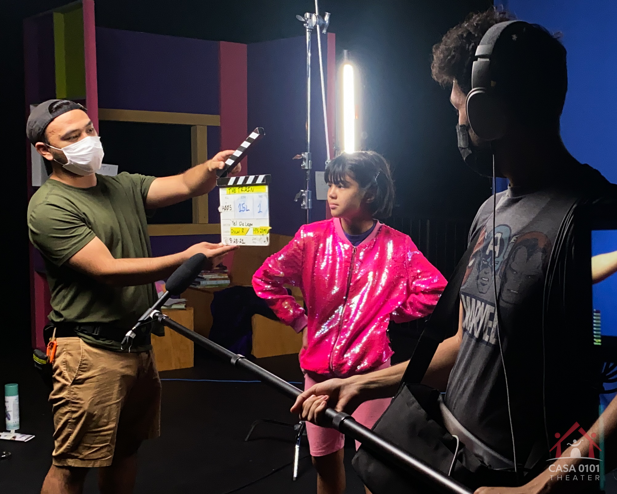 A production member holds a slate agains a student actor. A sound engineer holds a boom microphone up agains the student to record her sound.