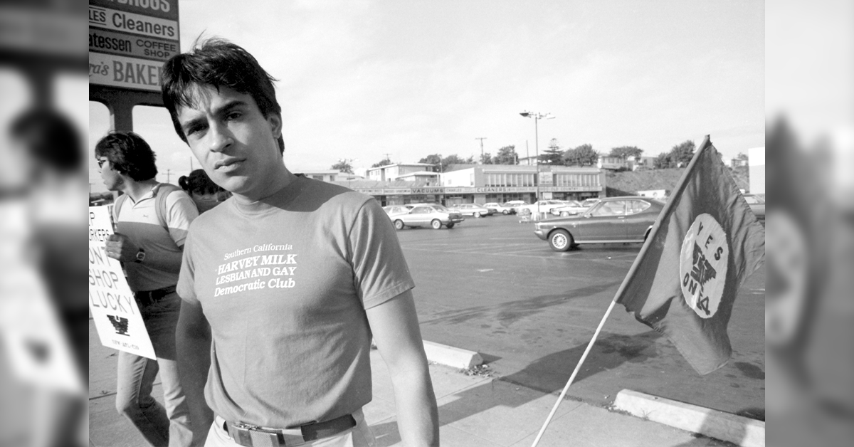 A black and white photo of a man, looking at the camera. He is standing in a parking lot in protest.