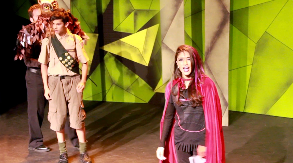 Production still of a show featuring a young woman in a red cape. Behind her to the left is a young boy scout. A man with an owl puppet stands behind him.
