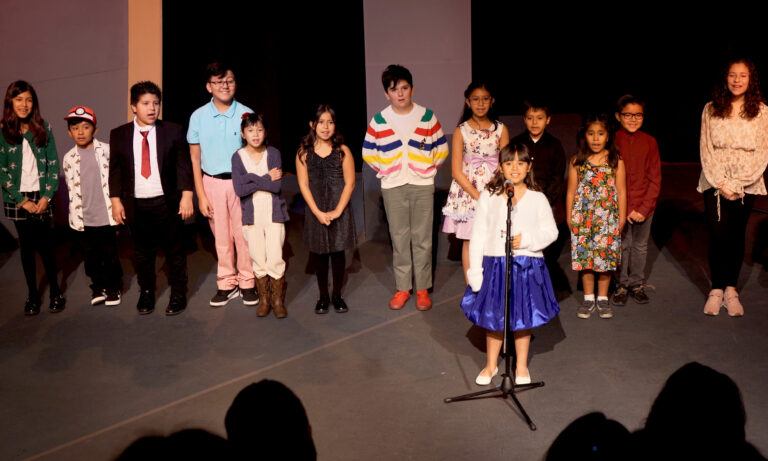 A group of school aged students are standing side by side on stage singing. A young girl is standing in front of them singing into a microphone.
